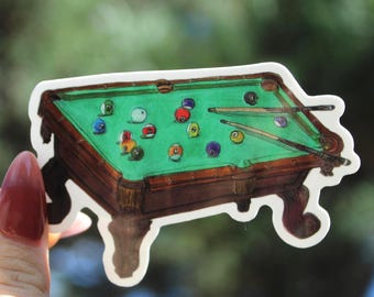 May include: A die-cut sticker featuring a brown wooden pool table with a green felt surface. The table is illustrated with various colored billiard balls scattered across the top and two wooden pool cues resting diagonally on the green surface.