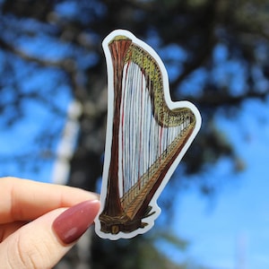 May include: A sticker featuring a detailed illustration of a harp. The harp is depicted with a brown wooden frame, gold accents, and numerous strings. The background is a blurred view of a blue sky and tree branches.