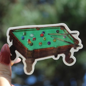 Op de afbeelding: Een sticker met een gedetailleerde illustratie van een pooltafel met groen vilt, een bruin houten frame en diverse gekleurde biljartballen. Twee keuen liggen op de tafel. De sticker heeft een witte rand.