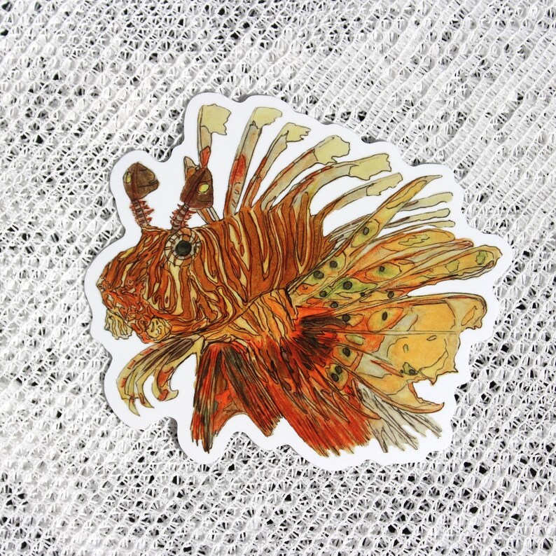 May include: A sticker featuring a detailed illustration of a lionfish. The lionfish is depicted in shades of orange, red, and yellow, with intricate fin details and a white border. The sticker is on a white, textured surface.
