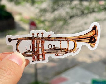 May include: A sticker depicting a brass trumpet, rendered in gold and brown tones. The illustration showcases the instrument's valves, tubing, and bell in detail. The sticker is held against a blurred outdoor backdrop, suggesting a musical theme.
