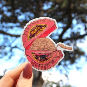 May include: A pink sticker of a Hubba Bubba bubble gum container, open to reveal a roll of gum. The container has the words "Hubba Bubba" in yellow and red. The sticker is held by a hand with a maroon nail polish.