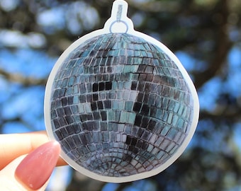 Op de afbeelding: Een sticker met een discobalontwerp. De bal is bedekt met kleine, vierkante, reflecterende tegels in blauwe en grijze tinten. Hij heeft een zilveren ring aan de bovenkant om op te hangen. De sticker wordt tegen een vage achtergrond van groen en blauw gehouden.
