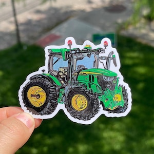 Könnte beinhalten: Ein Aufkleber mit einem grünen und gelben Traktor mit schwarzen Reifen. Der Traktor hat ein detailliertes Design, einschließlich Lichtern und einer Kabine. Der Aufkleber befindet sich auf weißem Hintergrund und scheint von einer Hand gehalten zu werden.
