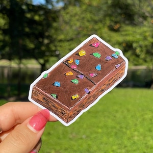 May include: A rectangular chocolate brownie sticker featuring a dark brown top surface decorated with small, multi-colored sprinkles in shades of blue, green, yellow, pink, and purple, with a textured brown side profile and a white die-cut border.