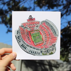 May include: A watercolour illustration of a stadium with red seating and a green field. The stadium is filled with red seating and has a green field with white markings. The top of the stadium has the word "CHAMPIONS".