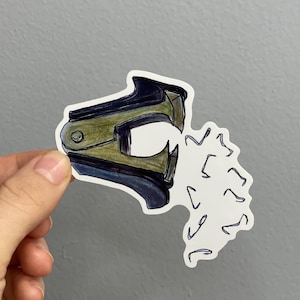 May include: A sticker featuring a hand-drawn illustration of a stapler expelling staples. The stapler is colored in shades of green, black, and blue. The staples are white with black outlines. The sticker is held against a gray background.