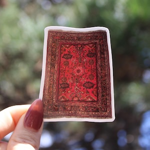 May include: A sticker featuring a detailed illustration of a Persian rug. The rug design is predominantly red with intricate patterns in shades of brown and gold. The sticker has a white border and is held against a blurred green background.