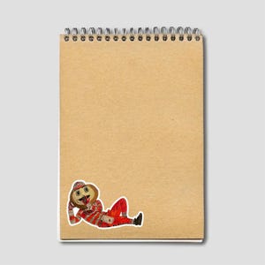 May include: A spiral-bound notepad with a tan cover. A sticker of a cartoon character in a red and white striped outfit, hat, and boots is affixed to the lower right corner of the notepad. The character is lying down and appears to be smiling.