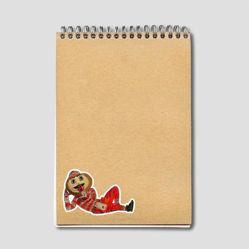 May include: A spiral-bound notepad with a tan cover. A sticker of a cartoon character in a red and white striped outfit, hat, and boots is affixed to the lower right corner of the notepad. The character is lying down and appears to be smiling.