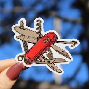 Op de afbeelding: Een sticker met een illustratie van een rood en bruin Zwitsers zakmes. Het mes heeft meerdere gereedschappen, waaronder een mes, schaar en een kurkentrekker. De rode handgreep heeft een wit embleem. De sticker wordt tegen een vage blauwe achtergrond gehouden.