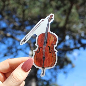 May include: A sticker featuring a cello with a bow. The cello is depicted in shades of brown and orange, with black strings and details. The bow is light brown. The sticker is held against a blurred background of green and blue.