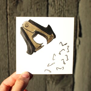 Peut inclure: Illustration à l'aquarelle d'un arrache-agrafes doré et noir sur un carré blanc. Plusieurs formes d'agrafes sont dessinées à droite de l'arrache-agrafes. L'image est tenue par une main.