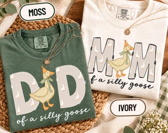 May include: A moss green t-shirt with the text DAD of a silly goose and an ivory t-shirt with the text MOM of a silly goose. Both shirts feature a graphic of a goose wearing a party hat and a green ribbon, with small goose illustrations on the letters.