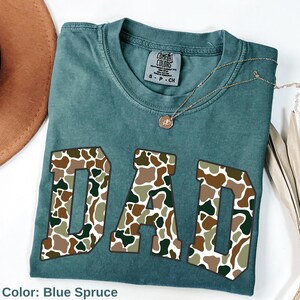 May include: Blue spruce t-shirt with the word "DAD" in large, camouflage-patterned letters. A gold necklace with a pendant rests on the shirt. The shirt is folded and displayed with a brown hat and dried plant stems.