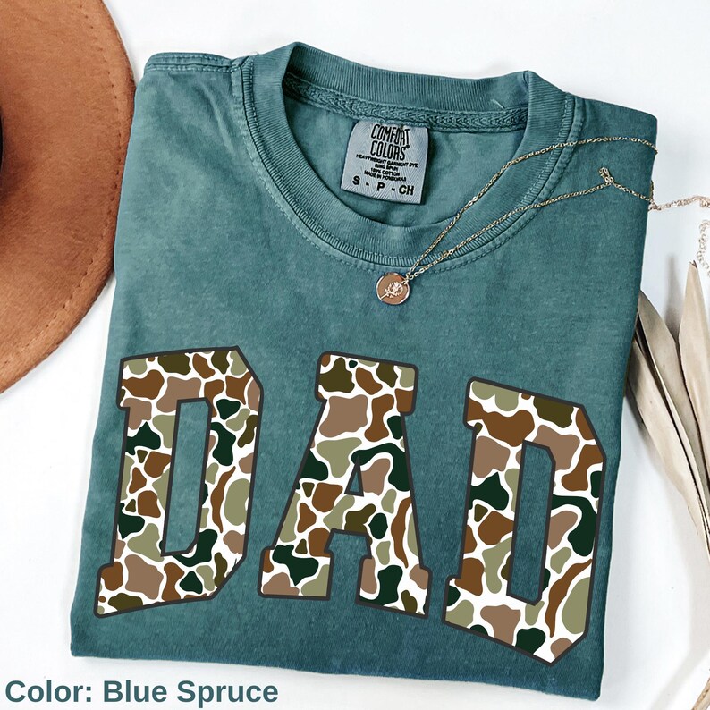 May include: Blue spruce t-shirt with the word "DAD" in large, camouflage-patterned letters. A gold necklace with a pendant rests on the shirt. The shirt is folded and displayed with a brown hat and dried plant stems.