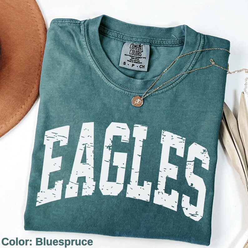 May include: A blue-green t-shirt with the word "EAGLES" in distressed white lettering. The shirt has a crew neck and a small tag at the neckline. A gold necklace with a pendant is draped across the shirt.
