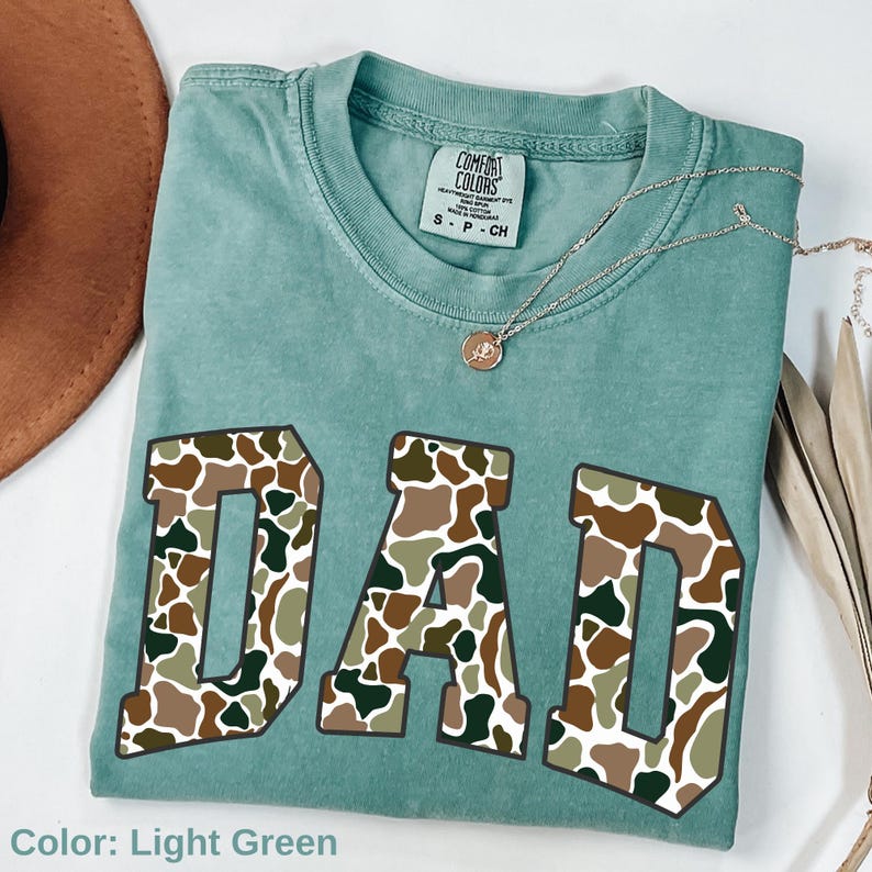 May include: Light green t-shirt with the word "DAD" in large, camouflage-patterned letters. The shirt has a crew neck and short sleeves. A silver necklace with a pendant is draped across the shirt.