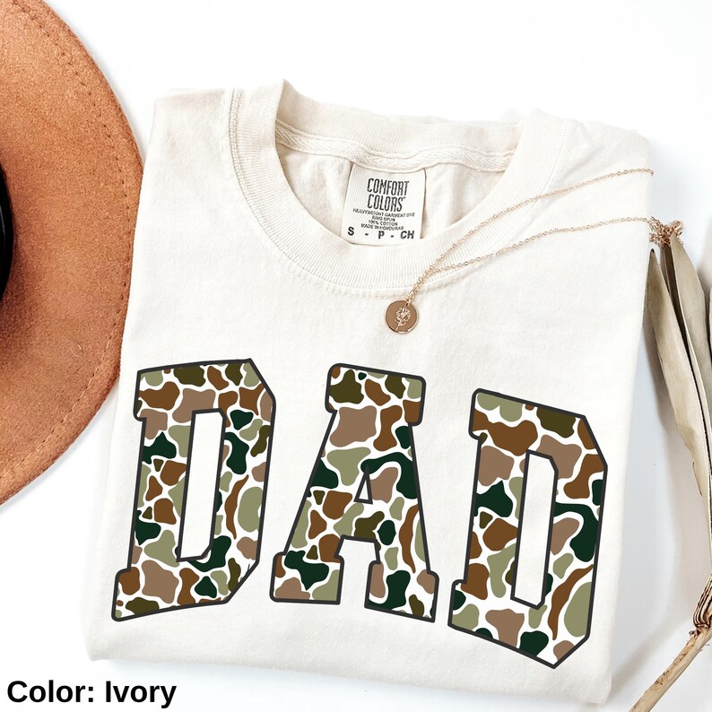 May include: Ivory-colored t-shirt with the word "DAD" in large, camouflage-patterned letters. The shirt has a crew neck and short sleeves. A gold necklace with a pendant is draped across the shirt.