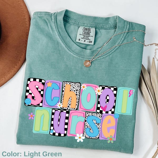 May include: Light green Comfort Colors t-shirt with the words "School Nurse" in colorful, block letters. The design includes daisies, stars, and a lightning bolt. A silver necklace with a pendant is draped across the shirt.