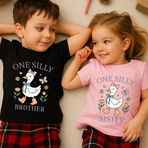 May include: Two children wearing matching t-shirts. One shirt is black with the words "ONE SILLY BROTHER" and a cartoon duck graphic. The other shirt is pink with the words "ONE SILLY SISTER" and a similar duck graphic.