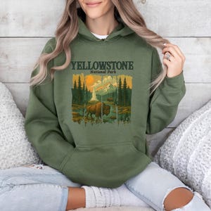 May include: Olive green hoodie featuring a graphic of Yellowstone National Park. The design includes a bison, mountains, trees, and the text "YELLOWSTONE National Park". The hoodie has a drawstring hood and a front pocket.
