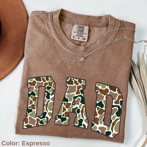 May include: Espresso-colored t-shirt with the word "DAD" in a camouflage pattern. The shirt has a crew neck and short sleeves. A silver necklace with a pendant is draped across the shirt.