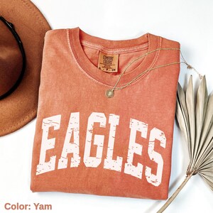 May include: A yam-colored Comfort Colors t-shirt with the word "EAGLES" in distressed white lettering. A gold necklace with a pendant is draped over the shirt. A brown hat and dried palm leaf are in the background.