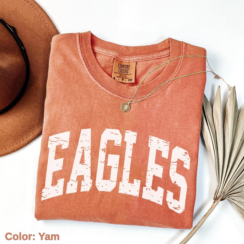 May include: A yam-colored Comfort Colors t-shirt with the word "EAGLES" in distressed white lettering. A gold necklace with a pendant is draped over the shirt. A brown hat and dried palm leaf are in the background.