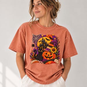 May include: A rust-colored short-sleeve t-shirt featuring a graphic print of a black cat, a jack-o'-lantern, a haunted house, a spider web, and a witch hat. The design includes a sunflower, bats, and a colorful patchwork pattern at the base.