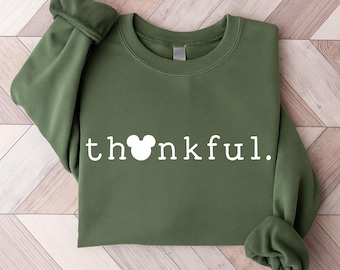 May include: An olive green sweatshirt with the word "thankful" printed in white. The "o" in the word is replaced with a Mickey Mouse head silhouette. The sleeves are rolled up. The sweatshirt is made of a soft material.