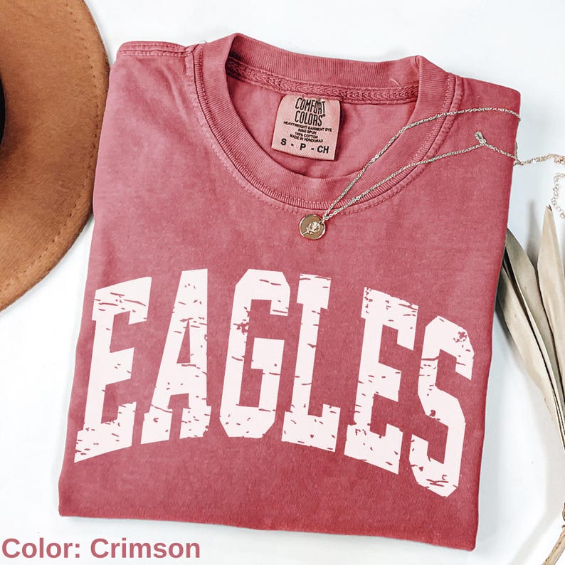 May include: Crimson-colored t-shirt with the word "EAGLES" in distressed white lettering. The shirt has a crew neck and short sleeves. A silver necklace with a round pendant is draped across the shirt. The shirt is made by Comfort Colors.