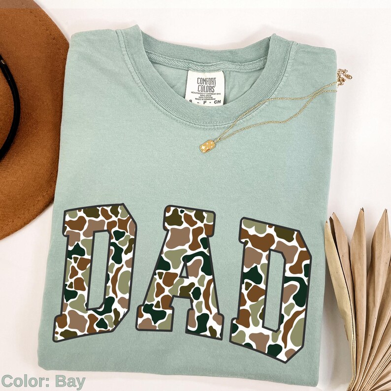 May include: A light green Comfort Colors t-shirt with the word "DAD" in large, block letters. The letters are filled with a camouflage pattern in shades of brown, green, and white. A gold necklace and a brown hat are also visible.