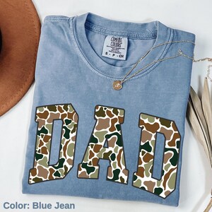 May include: Blue jean colored t-shirt with the word "DAD" in large, block letters. The letters are filled with a camouflage pattern in shades of brown, green, and tan. A gold necklace with a pendant rests on the shirt.