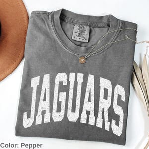 May include: A pepper-colored short-sleeve t-shirt with a crew neckline and a Comfort Colors label. The front features the word JAGUARS in large, distressed white block lettering. A delicate gold-toned chain necklace with a small round pendant rests on the fabric.
