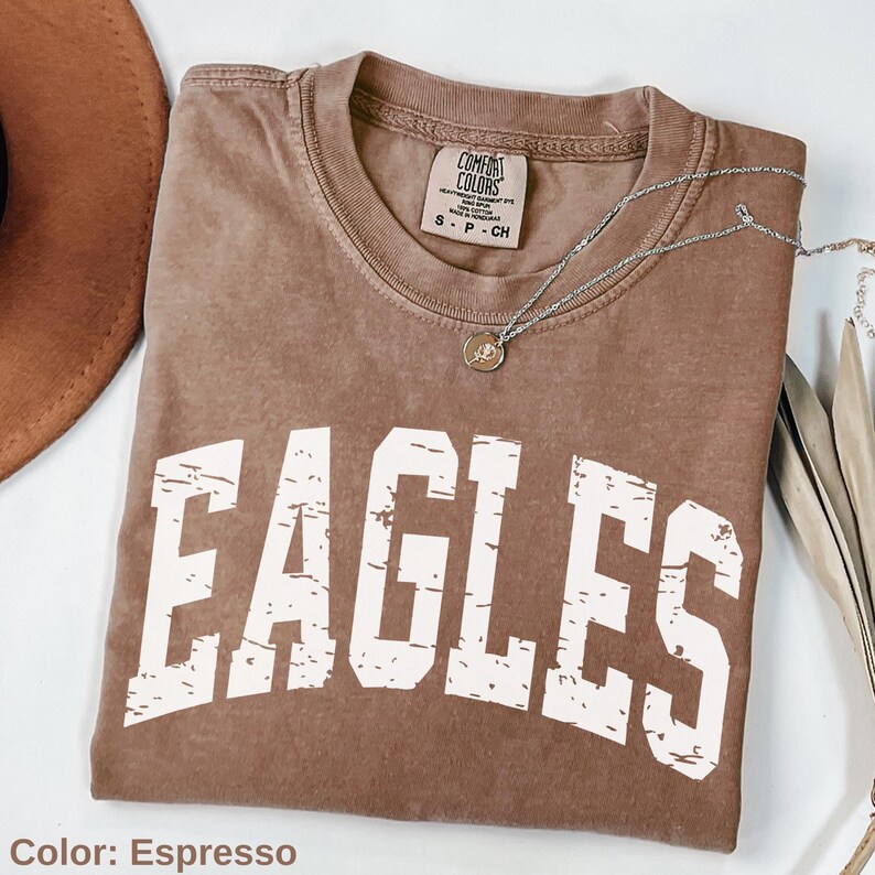 May include: Espresso-colored t-shirt with the word "EAGLES" in distressed white lettering. The shirt has a crew neck and a small tag at the neckline. A silver necklace with a pendant is draped across the shirt. A brown hat is visible in the upper left corner.