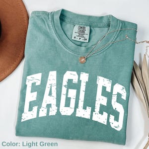May include: Light green t-shirt with the word "EAGLES" in distressed white lettering. The shirt has a crew neck and short sleeves. A silver necklace with a pendant is draped across the shirt. The shirt is folded on a white surface.