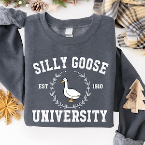May include: Dark gray sweatshirt with the text "SILLY GOOSE UNIVERSITY EST 1910" in white. A white goose illustration is centered within a laurel wreath. The sweatshirt is a casual, comfortable garment.