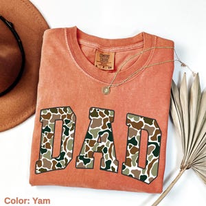 May include: A yam-colored Comfort Colors t-shirt with the word "DAD" in large, camouflage-patterned letters. A gold necklace with a pendant rests on the shirt. A brown hat and dried plant are in the background.