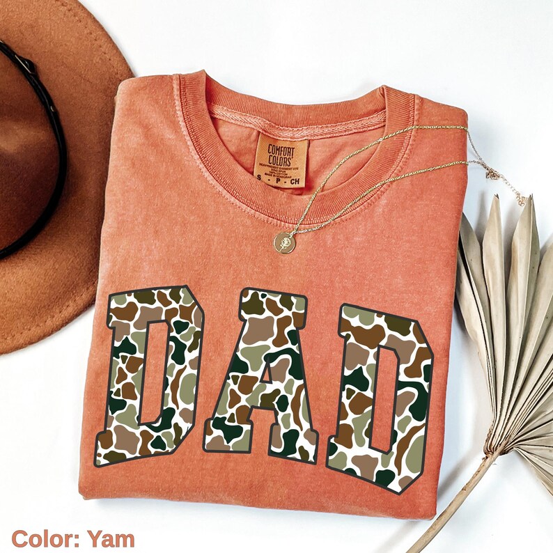 May include: A yam-colored Comfort Colors t-shirt with the word "DAD" in large, camouflage-patterned letters. A gold necklace with a pendant rests on the shirt. A brown hat and dried plant are in the background.