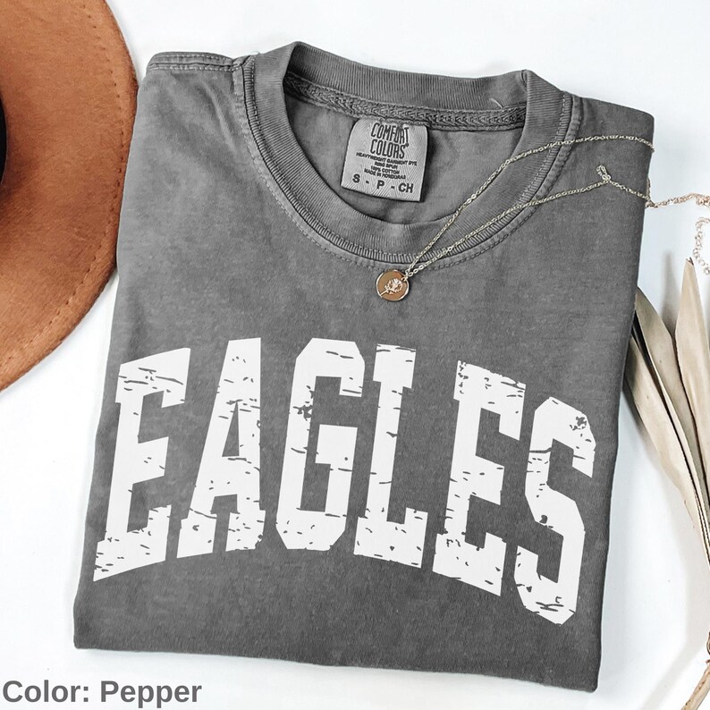May include: A gray Comfort Colors t-shirt with the word "EAGLES" in distressed white lettering. A silver necklace with a pendant is draped across the shirt. The shirt is folded on a white surface.