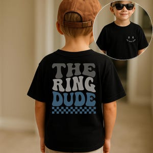 May include: Black t-shirt with the text "THE RING DUDE" in gray and blue, with a checkered pattern at the bottom. A small smiley face and the same text are on the front. The child is wearing a brown baseball cap.