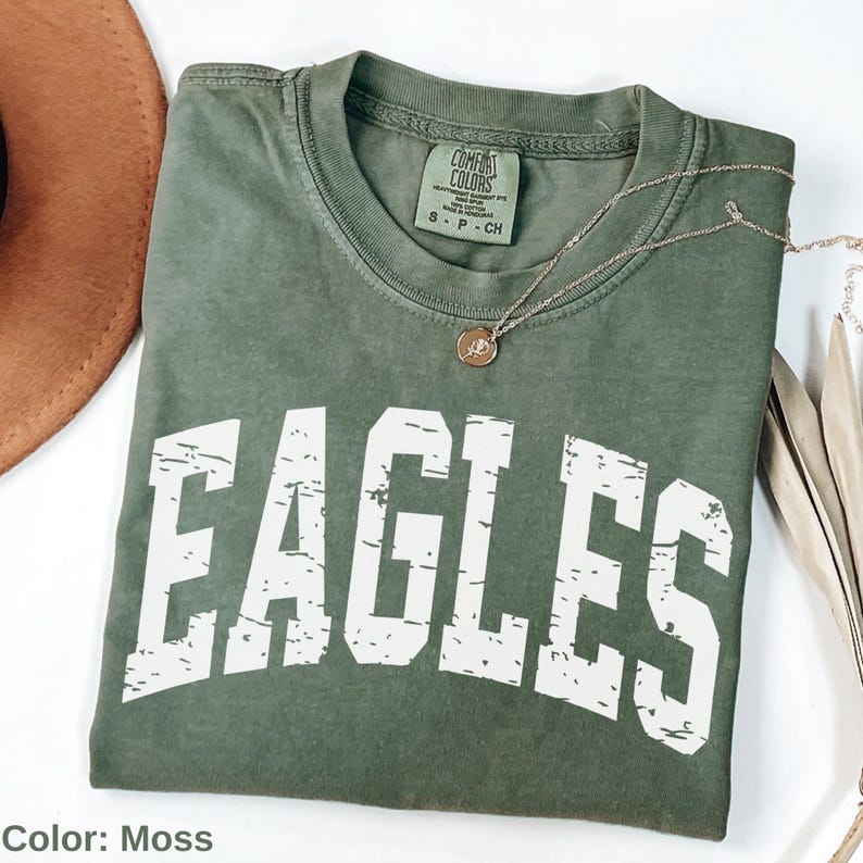 May include: A moss green t-shirt with the word "EAGLES" printed in distressed white lettering. The shirt is folded and has a Comfort Colors tag at the neckline. A gold necklace with a pendant is draped across the shirt.