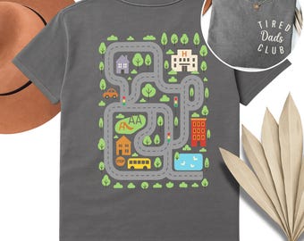 May include: A gray t-shirt with a colorful road map design on the back, featuring houses, trees, a bus, and a car. The front of the shirt has the text "Tired Dads Club".