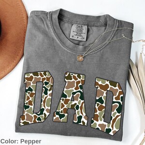 May include: A gray Comfort Colors t-shirt with the word "DAD" in a camouflage pattern. The shirt has a crew neck and short sleeves. A silver necklace with a small pendant is draped on the shirt. The color of the shirt is pepper.