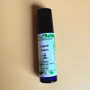 May include: A blue glass roller bottle with a black cap and a white label. The label features green watercolor accents and black text that reads Empowered Essentials, Liquid Xanax, Calming Aromatherapy Blend, 10 ML, and External Use Only.