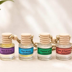 Puede incluir: Cuatro frascos de vidrio pequeños con tapas de madera clara y cuerdas para colgar. Cada frasco tiene una etiqueta de color con el texto: Warm & Cozy, Clear Airway, Empower-gize y Clean & Empowered, incluyendo la marca Empowered Essentials.