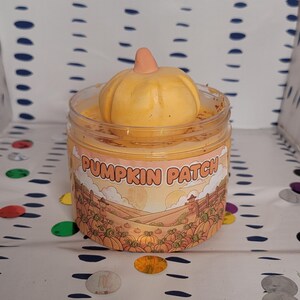 May include: A clear plastic jar filled with orange-colored slime, topped with a pumpkin-shaped decoration. The jar has a label that reads "PUMPKIN PATCH" with a fall-themed illustration. The background is a white surface with blue and white polka dots.