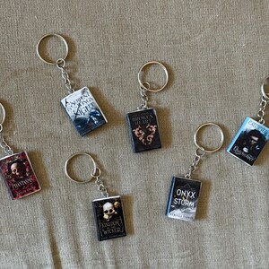 May include: Miniature book keychains with silver key rings. The keychains feature book covers with titles like "Serpent & Night", "Broken Heart", and "Onyx Storm". The keychains are on a tan fabric surface.