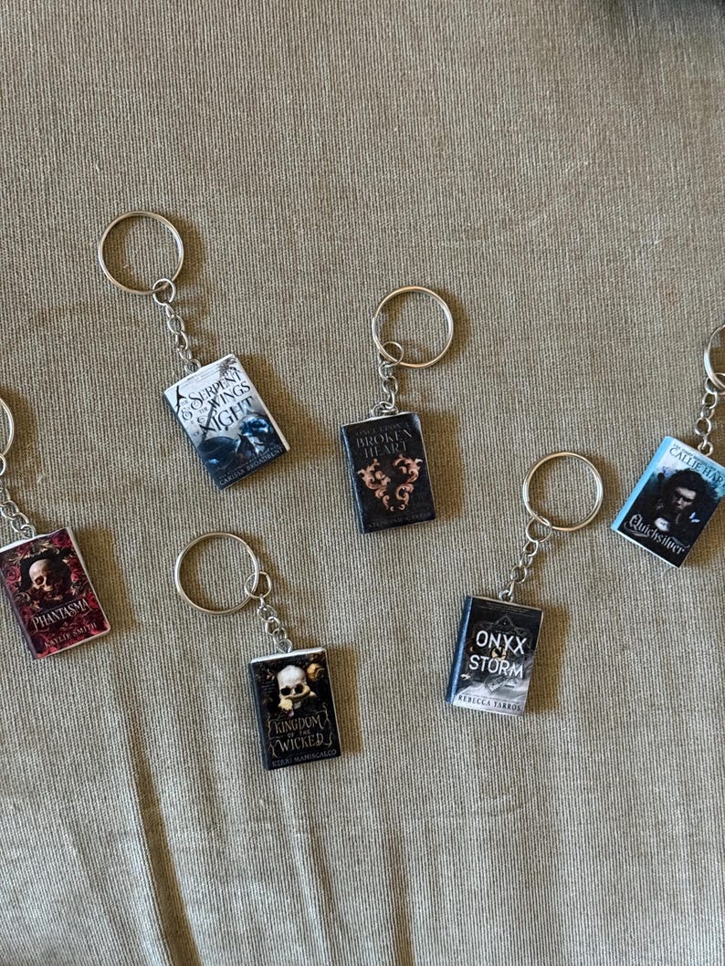 May include: Miniature book keychains with silver key rings. The keychains feature book covers with titles like "Serpent & Night", "Broken Heart", and "Onyx Storm". The keychains are on a tan fabric surface.
